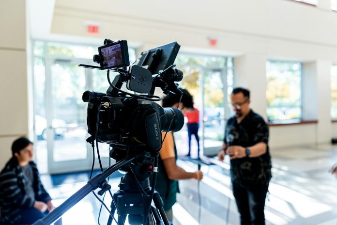 Videó riport: iparági konszolidáció — Close-up of a professional video camera on a tripod with people blurred in backg