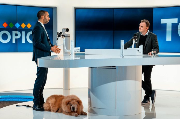 Vizuális magyarázó: kelet-európai kontextus — Two hosts discussing at a modern TV studio with a dog laying by. Engaging profes