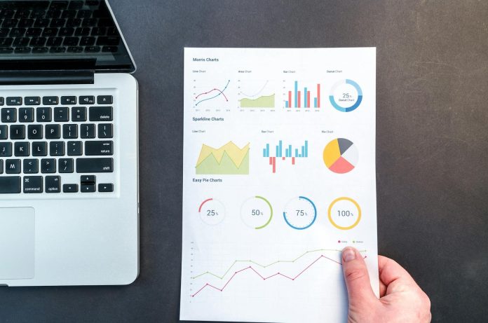 Infografika riport: iparági konszolidáció — Detailed charts and graphs on a document next to a laptop, representing data ana