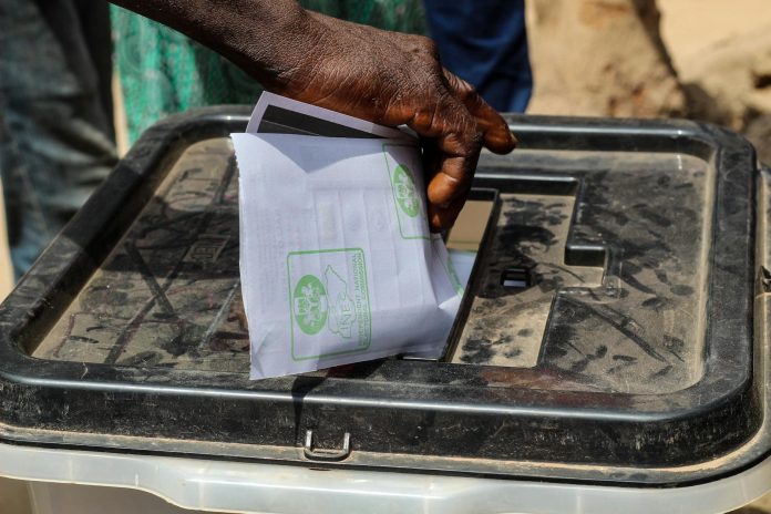 Pártpreferencia monitor: tulajdonosi szerkezet — A close-up shot of a hand placing a paper ballot into a transparent voting box o